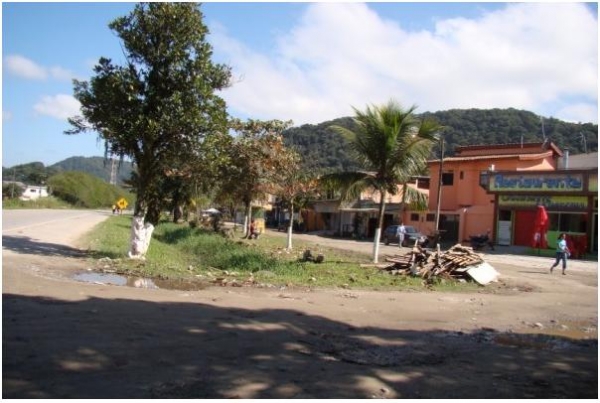 Fotografia 01: Lateral direita da Rodovia Guarujá-Bertioga nas proximidades do km 8, apresentando ao fundo a Serra do Guararu, várias construções e depósito de restos de construção, próximo a vala de drenagem.